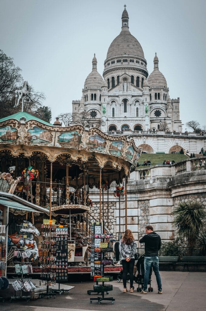 Montmartre Carousel & Sacré-Cœur – Paris with Kids The Montmartre Carousel with the Sacré-Cœur Basilica in the background on a cloudy day in Paris. A charming family-friendly attraction in Montmartre.